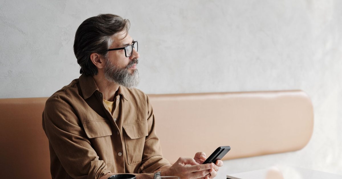 Bärtiger Mann im Café mit Handy in der Hand, nachdenklich zur Seite blickend