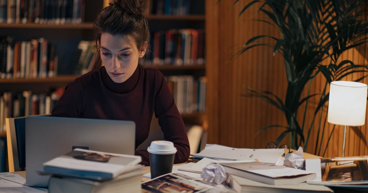 Frau arbeitet an einem Schreibtisch voller aufgeschlagener Bücher und einem Laptop